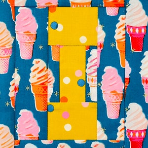 May include: A yellow fabric block with white polka dots and a pattern of ice cream cones on a blue background. The fabric block is divided into four sections, each with a different pattern.