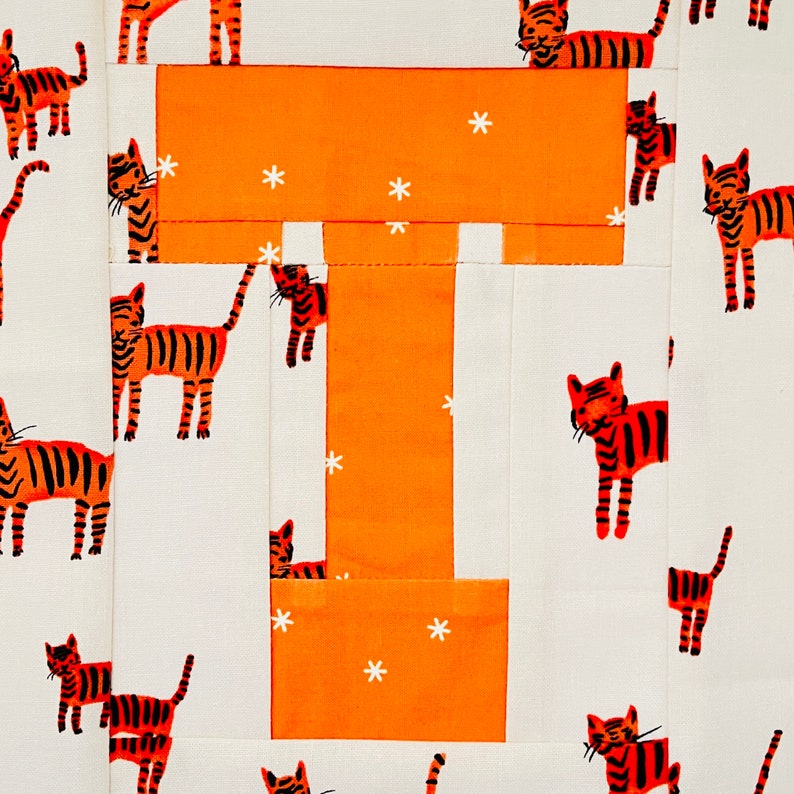 May include: A white fabric with a large orange letter T in the centre. The letter T is made up of smaller orange fabric squares. The fabric is decorated with small orange and white tiger illustrations.