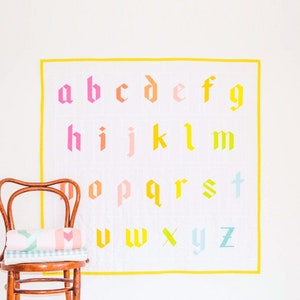 Puede incluir: Una colcha blanca con un borde amarillo que presenta el alfabeto en letras de estilo gótico de colores. Una silla de madera con una colcha enrollada en el asiento se encuentra delante de la colcha.