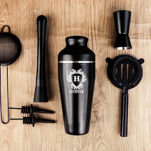 May include: A black cocktail shaker set with a variety of bar tools, including a strainer, muddler, jigger, and bar spoon. The shaker is engraved with a monogram and the name "Hunter". The set is arranged on a wooden surface.