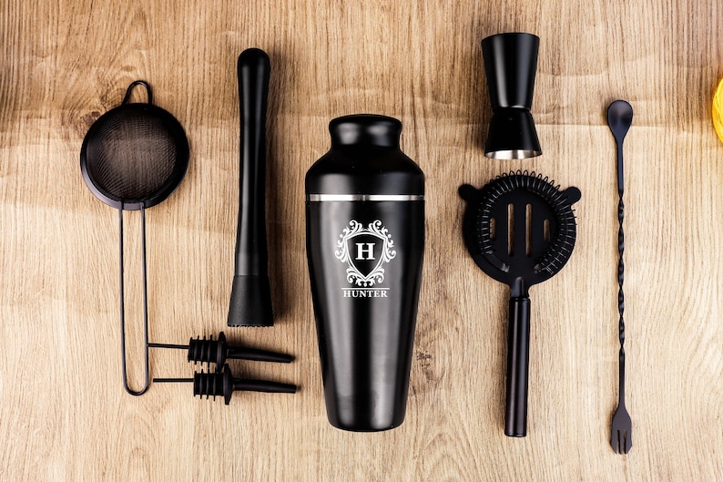 May include: A black cocktail shaker set with a variety of bar tools, including a strainer, muddler, jigger, and bar spoon. The shaker is engraved with a monogram and the name "Hunter". The set is arranged on a wooden surface.