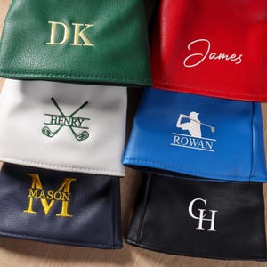 Könnte beinhalten: Eine Sammlung von sechs Golfschlägerhauben aus Leder in Grün, Rot, Weiß, Blau, Marineblau und Schwarz. Jede Haube ist mit individuellen Stickereien versehen, darunter Initialen wie DK, GH, MASON oder Namen wie James, Henry und Rowan mit Golfmotiven.
