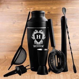 Puede incluir: Un juego de coctelera negro con un diseño de escudo personalizado que incluye la letra "H" y el nombre "HUNTER". El juego incluye una coctelera, un colador, un jigger, un mortero, una cuchara de bar y boquillas. Las herramientas son negras.