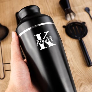May include: A black cocktail shaker with a silver band and the name "KIRSTY" in white lettering. Cocktail tools, including a strainer, jigger, and bar spoon, are arranged on a wooden surface.