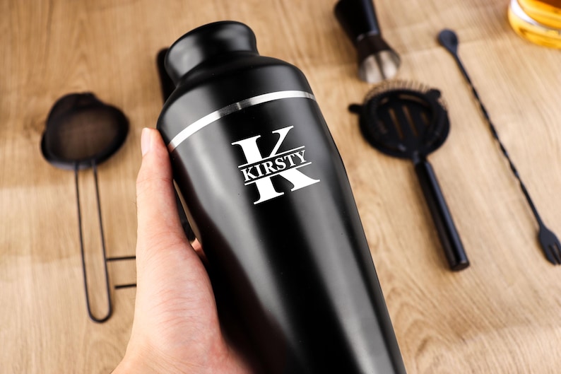 May include: A black cocktail shaker with a silver band and the name "KIRSTY" in white lettering. Cocktail tools, including a strainer, jigger, and bar spoon, are arranged on a wooden surface.