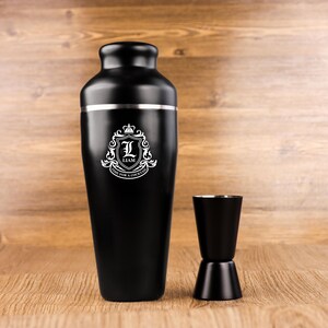 May include: Black cocktail shaker with a silver band and a matching black jigger. The shaker features a white crest with the letter "L" and the name "Liam". The text "TIME FOR A COCKTAIL" is also on the crest.