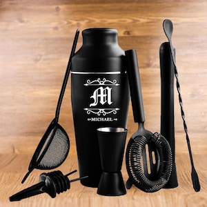 May include: A black cocktail shaker set with a personalized design. The set includes a shaker, strainer, jigger, muddler, bar spoon, and pour spouts. The shaker is engraved with the letter "M" and the name "MICHAEL".