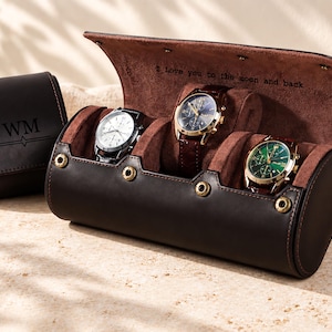 May include: A dark brown leather watch roll case with gold-tone snap buttons, holding three wristwatches. The interior is lined with brown suede and features the engraved text I love you to the moon and back. A matching watch case with the initials WM is nearby.