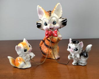 May include: Three ceramic cat figurines, one orange and white tabby cat with a red bow tie and two smaller cats, one orange and white and one black and white, connected by a gold chain.