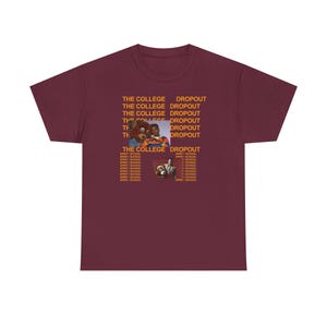 Puede incluir: Camiseta burdeos con el texto "THE COLLEGE DROPOUT" repetido. El diseño incluye imágenes de un oso de peluche y un hombre con traje. Es una prenda de vestir informal.