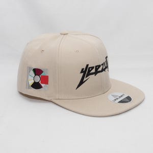 Könnte beinhalten: Eine beige Baseballkappe mit einem schwarzen "YEEZY"-Logo, das auf der Vorderseite gestickt ist, und einem bunten Patch an der Seite. Die Kappe hat ein schwarzes "UNLIMIT"-Etikett auf der Rückseite.