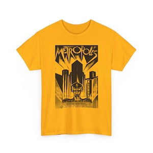 Puede incluir: Camiseta amarillo mostaza con un gráfico negro y marrón de la película "Metrópolis". El gráfico incluye el título de la película, un paisaje urbano futurista y una figura de robot. También se ve el texto "Ein Film von Fritz Lang".