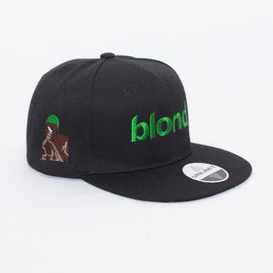 Op de afbeelding: Zwarte baseballpet met een groen "blonded" logo en een bruine en groene afbeelding aan de zijkant. De pet heeft een zwarte verstelbare band met een "UNLIMIT" label.