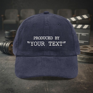 Puede incluir: Una gorra de béisbol de pana azul marino con texto bordado en blanco en la parte delantera que dice PRODUCED BY "YOUR TEXT". La gorra tiene una corona estructurada, una visera curva y un botón en la parte superior.