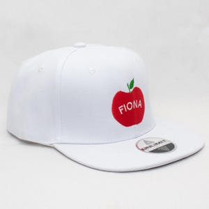 Puede incluir: Gorra de béisbol blanca con un diseño de manzana roja y el nombre "FIONA". La gorra tiene una visera plana y una pegatina en la visera. Es un estilo clásico.