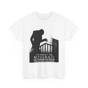 Puede incluir: Camiseta blanca con un gráfico en blanco y negro de la película "Nosferatu" de 1922. El diseño incluye una silueta del vampiro, el título de la película y el nombre del director. La camiseta es de material suave.