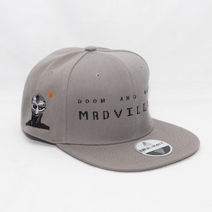 Può includere: Un cappello da baseball grigio con il testo "DOOM AND MADVILLAIN" ricamato sul davanti. Il cappello ha un'immagine in bianco e nero di un viso umano sul lato sinistro. Il cappello ha un logo nero "UNLIMIT" sul retro.