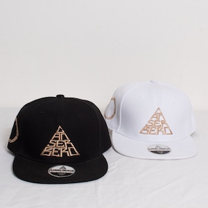 Può includere: Due cappellini da baseball, uno nero e uno bianco, sono esposti. Entrambi i cappellini presentano testo ricamato in oro e un design triangolare. Il cappellino nero ha una visiera nera, mentre quello bianco ha una visiera bianca. I cappellini sono su una superficie bianca.