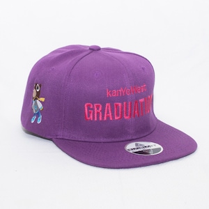 Könnte beinhalten: Lilafarbene Baseballkappe mit einem rosa gestickten Logo, auf dem "kanye west graduation" steht. Die Kappe hat einen kleinen gestickten Comic-Charakter an der Seite.