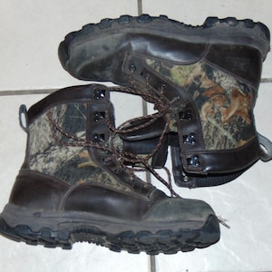 May include: A pair of brown leather hunting boots with camouflage print. The boots have a lace-up closure and a thick rubber sole.