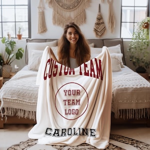 Peut inclure: Une couverture en polaire blanche avec le texte "Custom Team" en marron, un cercle avec le texte "Your Team Logo" en marron et le nom "Caroline" en noir.