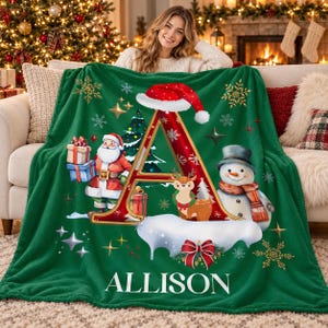 May include: A green fleece blanket featuring a large red letter A topped with a Santa hat. The design includes a cartoon Santa Claus holding gifts, a reindeer, a snowman with a scarf, and snowflakes. The name ALLISON is printed in white.