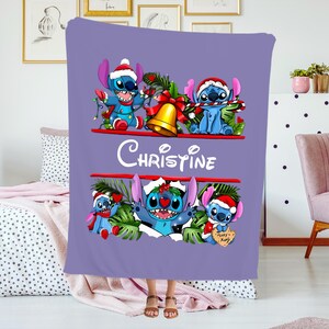 May include: A lilac blanket with Stitch characters wearing Santa hats and festive decorations. The blanket has the name "Christine" in white script. The design includes a gold bell and festive greenery, ideal for Christmas.