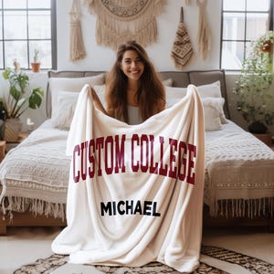 Peut inclure: Une couverture en polaire blanche avec le texte "CUSTOM COLLEGE" en lettres marron et le nom "MICHAEL" en lettres noires.