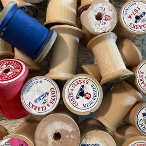 May include: A collection of vintage wooden spools in various sizes and colors. Some spools are natural wood, while others are painted red or blue. Several spools have labels with text, including "Coats & Clark's" and "Lark's".