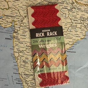 Puede incluir: Un paquete de cinta de festón roja mediana, 2,30 m de largo, con la marca "Wrights" y el color "Scarlet 76". La cinta está empaquetada sobre un mapa de la India.