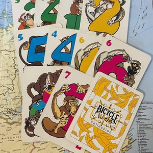 May include: A collection of Bicycle Kids playing cards featuring cartoon monkeys holding numbers 2 through 7, 10, and 3 through 6. The card backs have a yellow banana design with the words "Bicycle Kids collection".