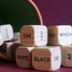 May include: Wooden dice with various words printed on them, including "BAD", "WHITE", "BLACK", and "ONE". The dice are spilling out of a brown container. The background is a dark red color.