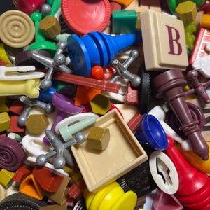 May include: A close-up of a collection of colorful game pieces. The image includes red, blue, yellow, green, and brown pieces. There are jacks, Scrabble tiles, and other game components. The pieces are scattered and overlapping.