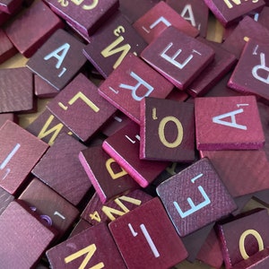 May include: A close-up of a pile of burgundy colored wooden letter tiles with gold and white lettering. The tiles are from a Scrabble game.