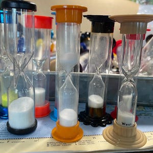 May include: A collection of colorful hourglasses with clear glass bodies and colored tops and bases. The sand timers have black, orange, red, and blue accents. One hourglass has the word "West" printed on it.