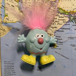 May include: A light blue, round-bodied toy figure with a pink feathered head, black eyes, and a red nose. It has yellow shoes and outstretched arms. The toy is placed on a map.