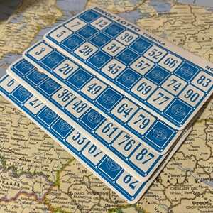 May include: A stack of blue and white Bingo Lotto Tombola cards with numbered squares. The cards are arranged on a vintage map, with the text "BINGO LOTTO TOMBOLA" visible at the top.