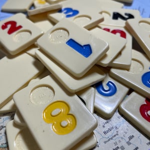 Puede incluir: Fichas de color marfil de un juego de Rummikub, con números en rojo, azul, amarillo y negro. Las fichas están esparcidas sobre un mapa, con los números 1, 2, 3 y 8 claramente visibles. Las piezas del juego tienen una textura ligeramente en relieve.