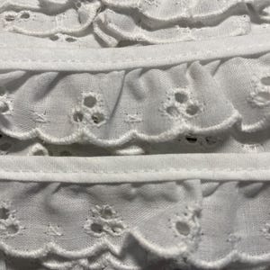 May include: Close-up of white eyelet fabric with scalloped edges and embroidered floral patterns. The fabric is layered, creating a textured, ruffled effect. The design features small, circular cutouts.