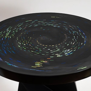 May include: A round black table with a smooth, glossy surface featuring a swirling, iridescent pattern of small, light-reflecting dashes that create a circular motion effect across the top, supported by a dark, solid, twisted pedestal base.