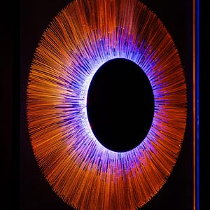 May include: A circular wall art piece featuring a central black void surrounded by a dense radial pattern of thin, orange-toned filaments. The inner edge of the circle is illuminated with a vibrant blue light that transitions into the orange rays.