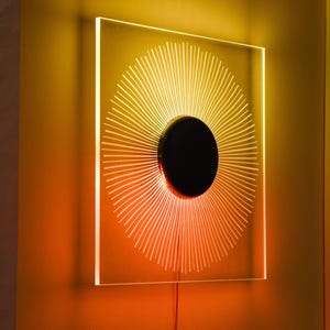 May include: A square, transparent acrylic wall light featuring a central black circular disc. The light emits a radiant, sunburst-like pattern of thin, glowing lines that transition from a bright yellow at the center to a deep orange at the edges.