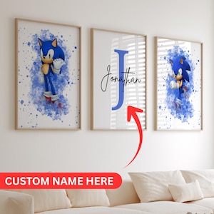 以下が含まれることがあります： 3つのフレーム入りプリント。2つは、青い水彩画のスプラッシュの背景にソニック・ザ・ヘッジホッグをフィーチャーしています。中央のプリントには、青い「J」が付いた「Jonathan」という名前が表示されています。画像の下部には「CUSTOM NAME HERE」というテキストがあります。