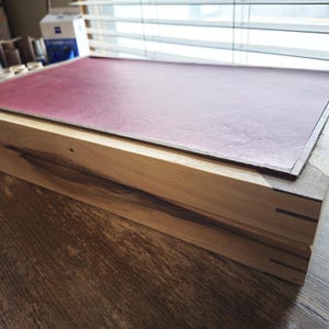 May include: A wooden desk with a burgundy leather top. The desk is made of light-colored wood with visible grain and a rectangular shape. The leather top is a solid color and appears to be smooth.