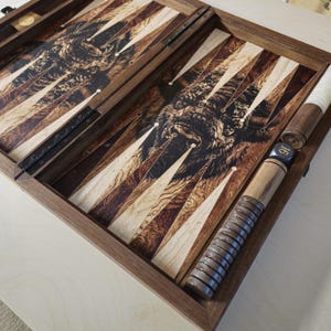 May include: A wooden backgammon set with a detailed bison illustration on the playing surface. The set is open, revealing the game board with alternating light and dark triangular points. A wooden dice cup and dice are visible.