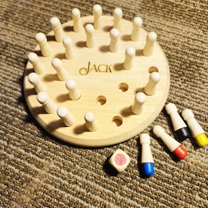 May include: A wooden game board with the word "JACK" engraved on it. The board has a circular design with pegs and holes. Several colored pegs and a die are next to the board. The game is on a brown surface.