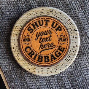 May include: A circular wooden cribbage board featuring a central tan leather inlay with the black text SHUT UP AND PLAY CRIBBAGE and the placeholder text your text here. The board has drilled holes for pegs and includes several metal game pegs.