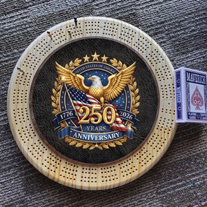 May include: A wooden cribbage board with a central design commemorating the 250th anniversary of the United States. The design features a golden eagle, American flag, and the text "1776-2026 250 Years Anniversary." A deck of Maverick playing cards is next to the board.