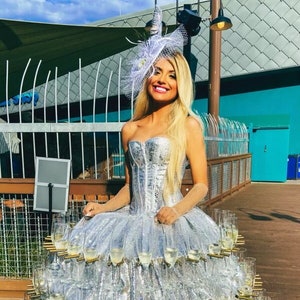 May include: A woman wearing a silver sequined dress with a champagne glass skirt. The skirt is made up of dozens of champagne glasses stacked on top of each other. The woman is wearing a white hat with a feather and has blonde hair. The background is a blue wall with a wooden railing.