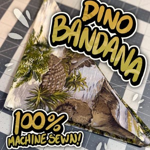 Könnte beinhalten: Dreieckiges Halstuch mit Dinosaurier-Print in Braun-, Grün- und Grautönen. Der Text "Dino Bandana" und "100% Machine Sewn!" sind in goldfarbenen Buchstaben dargestellt.